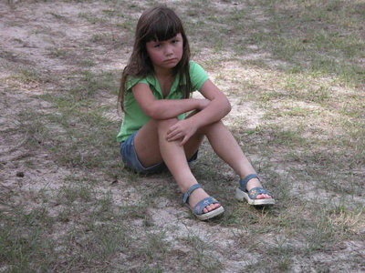 Primary age girl in t-shirt, shorts and sandals sitting on grass in thought, looking melancholy.